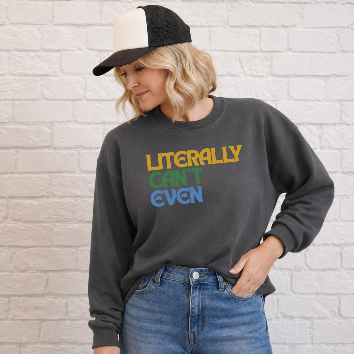 A woman wearing a dark charcoal gray sweatshirt with the text 'LITERALLY CAN'T EVEN' printed in yellow, green, and blue, paired with blue jeans and a black and white trucker hat, standing against a white brick wall.