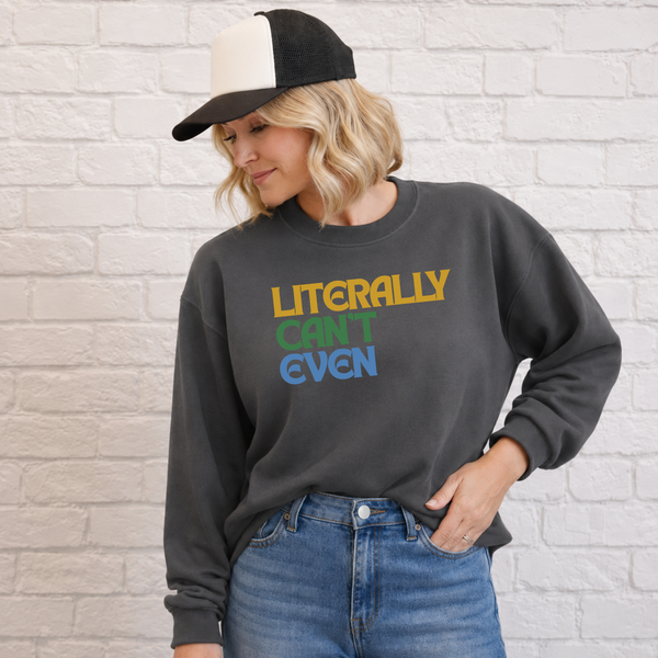 A woman wearing a dark charcoal gray sweatshirt with the text 'LITERALLY CAN'T EVEN' printed in yellow, green, and blue, paired with blue jeans and a black and white trucker hat, standing against a white brick wall.