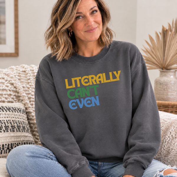 A woman sitting on a couch wearing a dark gray crewneck sweatshirt with the text 'LITERALLY CAN'T EVEN' printed on the chest in yellow, green, and blue.