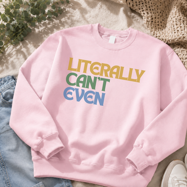A light pink crewneck sweatshirt with the text 'LITERALLY CAN'T EVEN' printed on the chest in yellow, green, and blue. The sweatshirt is styled in a flat-lay arrangement with light blue jeans, a white sneaker, and a beige knit blanket.