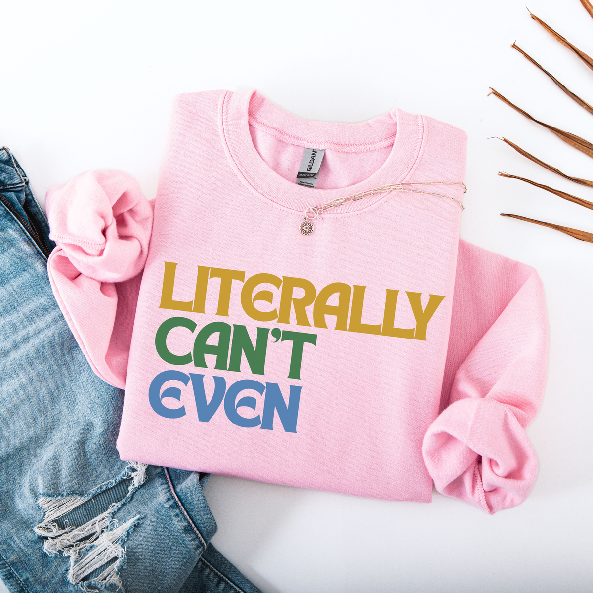 A folded light pink sweatshirt with rolled sleeves, featuring the text 'LITERALLY CAN'T EVEN' in yellow, green, and blue block letters. It is styled on a white background with a denim scrunchie and ripped blue jeans.