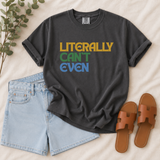 A dark charcoal gray graphic t-shirt with the text 'LITERALLY CAN'T EVEN' in yellow, green, and blue, styled in a flat lay with light blue denim shorts, brown slide sandals, and green eucalyptus branches on a beige fabric background.