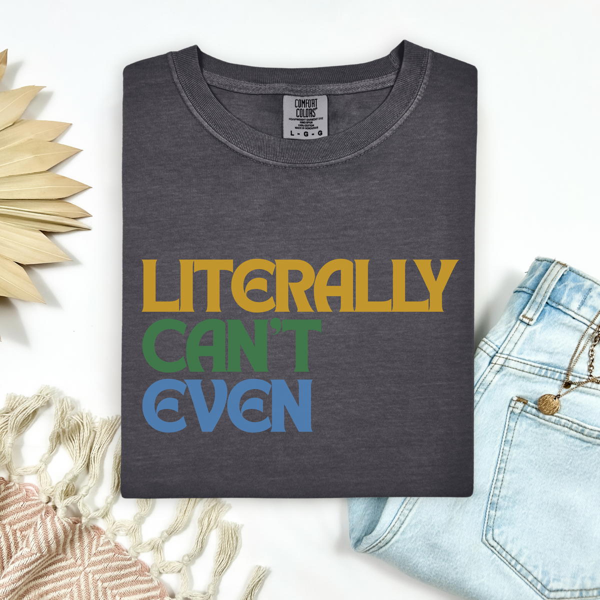 A folded dark gray graphic T-shirt with the phrase 'LITERALLY CAN'T EVEN' in yellow, green, and blue retro lettering, styled in a flat lay with light blue jeans, a woven textile, and a decorative sun-shaped object on a white background.
