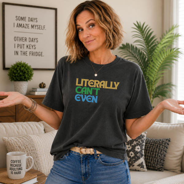 A woman wearing a dark charcoal graphic t-shirt with the text 'LITERALLY CAN'T EVEN' in yellow, green, and blue, paired with blue jeans in a cozy living room setting.