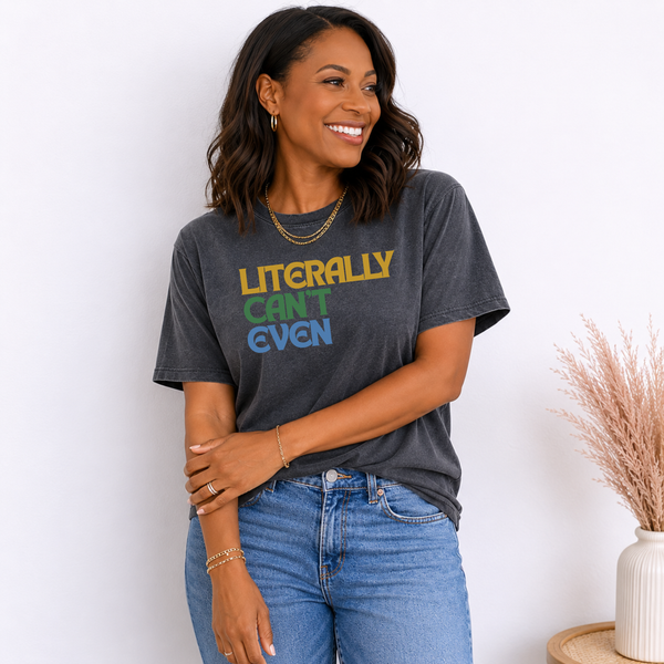 A woman wearing a dark gray graphic t-shirt with the text 'LITERALLY CAN'T EVEN' in yellow, green, and blue, paired with blue jeans, standing against a white wall.