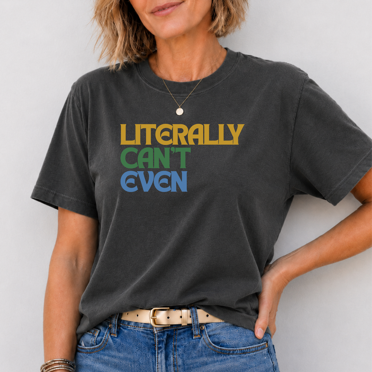 A woman wearing a dark charcoal gray graphic t-shirt with the text 'LITERALLY CAN'T EVEN' in gold, green, and blue. She is also wearing blue jeans and a light tan belt, standing against a plain light gray wall.