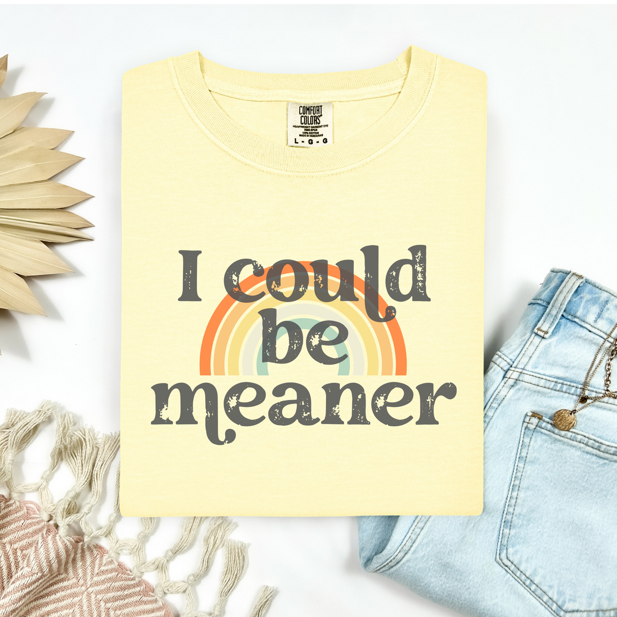 Yellow t-shirt with 'I could be meaner' text and rainbow graphic, placed on a white surface with decorative elements.