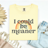 Yellow t-shirt with 'I could be meaner' text and rainbow graphic, placed on a white surface with decorative elements.