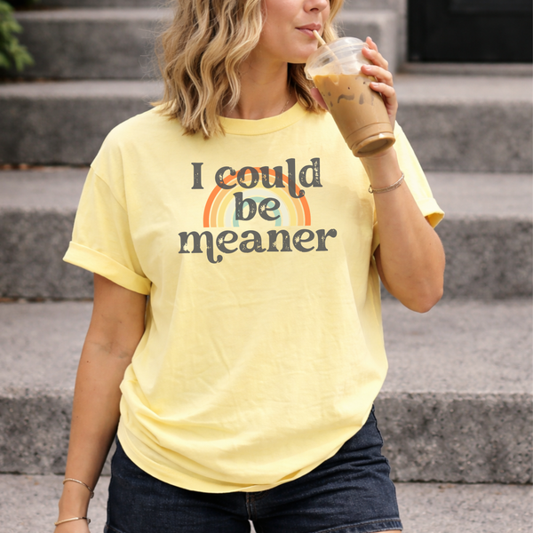 A woman wearing a yellow t-shirt with "I could be meaner: text, holding a drink outdoors.