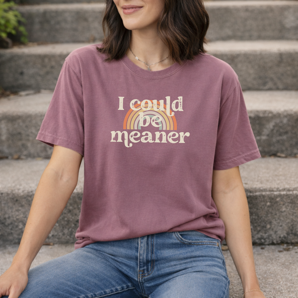 Woman wearing a berry pink purple t-shirt with a rainbow and text design, that says "I could be meaner" sitting on outdoor steps.