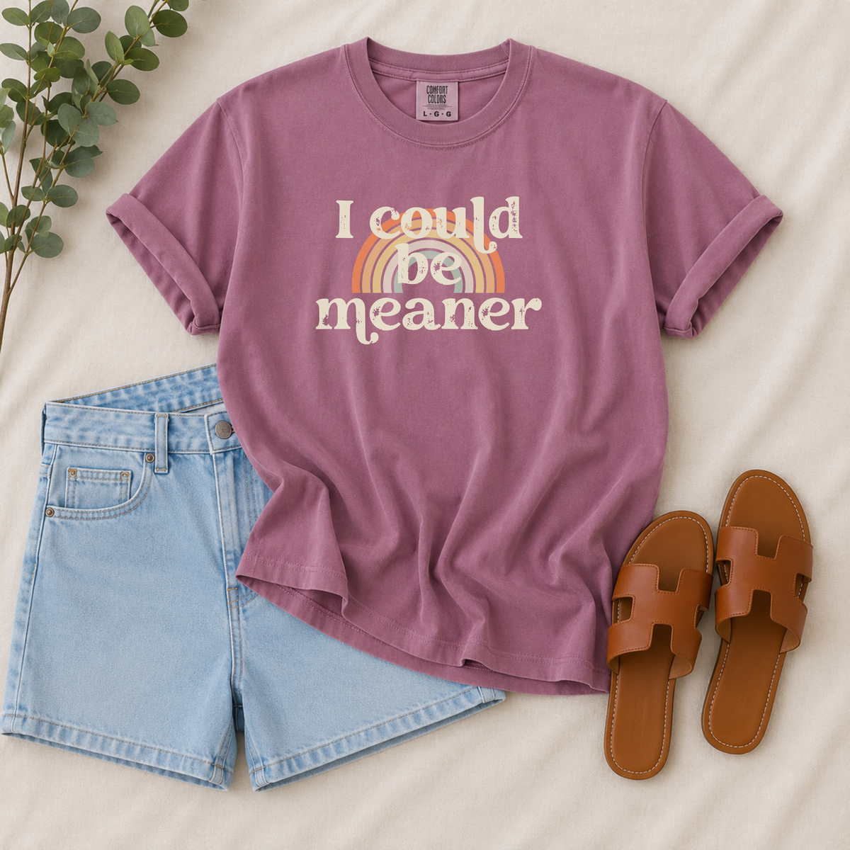 A mauve-purple graphic t-shirt with the text 'I could be meaner' and a retro rainbow design, styled in a flat lay with light-wash denim shorts, brown leather slide sandals, and a sprig of green leaves on a cream-colored fabric background.
