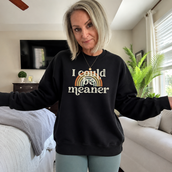 Woman wearing a black sweatshirt with a humorous graphic that says 'I could be meaner' and a rainbow in a living room.