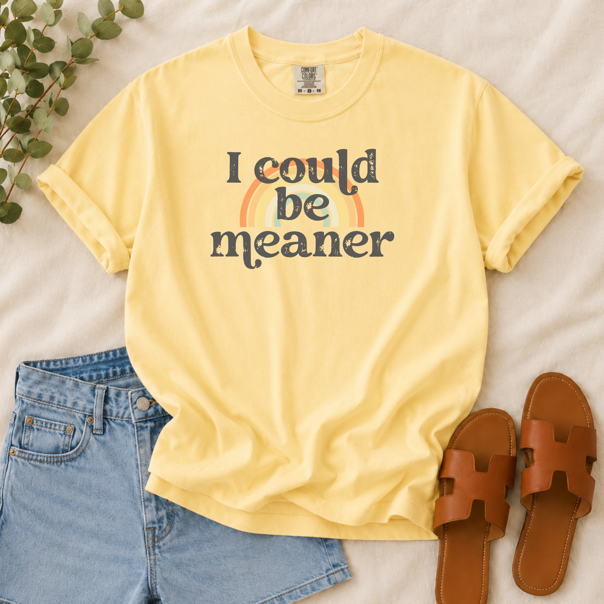 A pale yellow graphic T-shirt laid flat on a light fabric surface, styled with blue jeans, brown slide sandals, and a small green plant. The shirt has rolled sleeves and a front graphic reading 'I could be meaner' with a small rainbow behind the text.