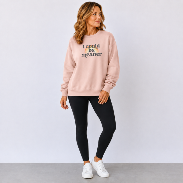 Woman wearing a pink sweatshirt with text 'i could be meaner' text over a rainbow and black leggings, and white sneakers on a light gray background.