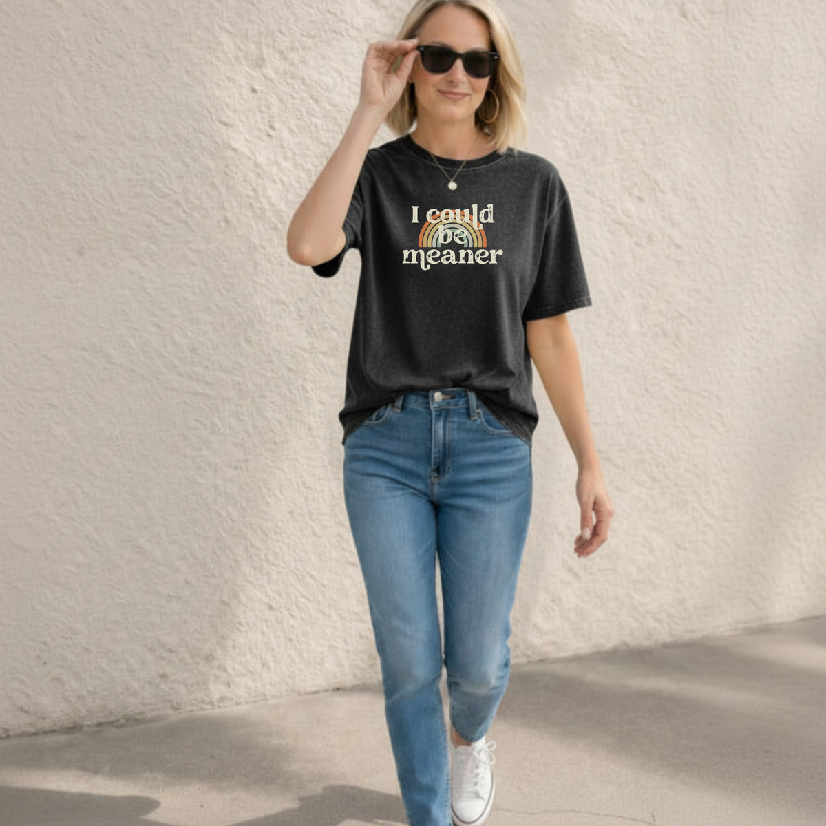 Woman wearing a faded pepper black t-shirt with a rainbow and text design, that says "I could be meaner" and jeans and sneakers walking outside.