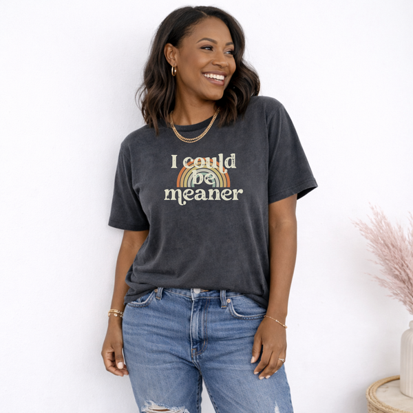 Woman wearing a faded pepper black t-shirt with a rainbow and text design, that says "I could be meaner" standing against a white background.