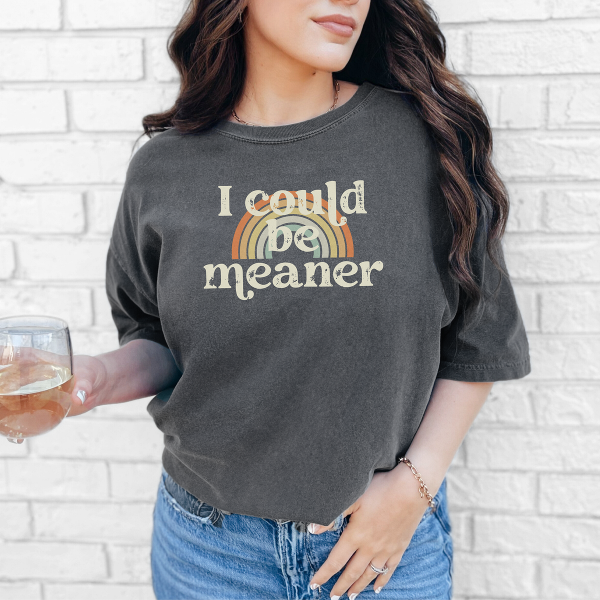 A woman wearing a dark gray t-shirt with 'I could be meaner' text and rainbow design, holding a glass of wine against a white brick wall.