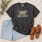 A dark charcoal gray graphic t-shirt with the text 'I could be meaner' and a retro rainbow design, styled in a flat lay with light blue denim shorts, brown leather slide sandals, and green eucalyptus leaves on a beige fabric background.