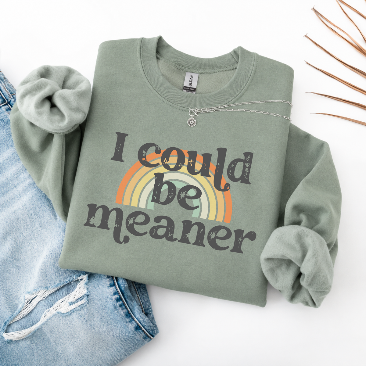 A folded green sweatshirt with 'i could be meaner' text over a rainbow and blue jeans on a white background.