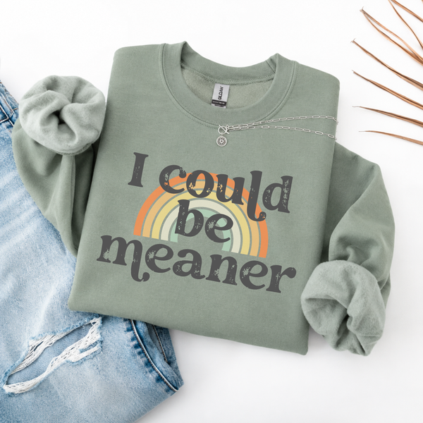 A folded green sweatshirt with 'i could be meaner' text over a rainbow and blue jeans on a white background.
