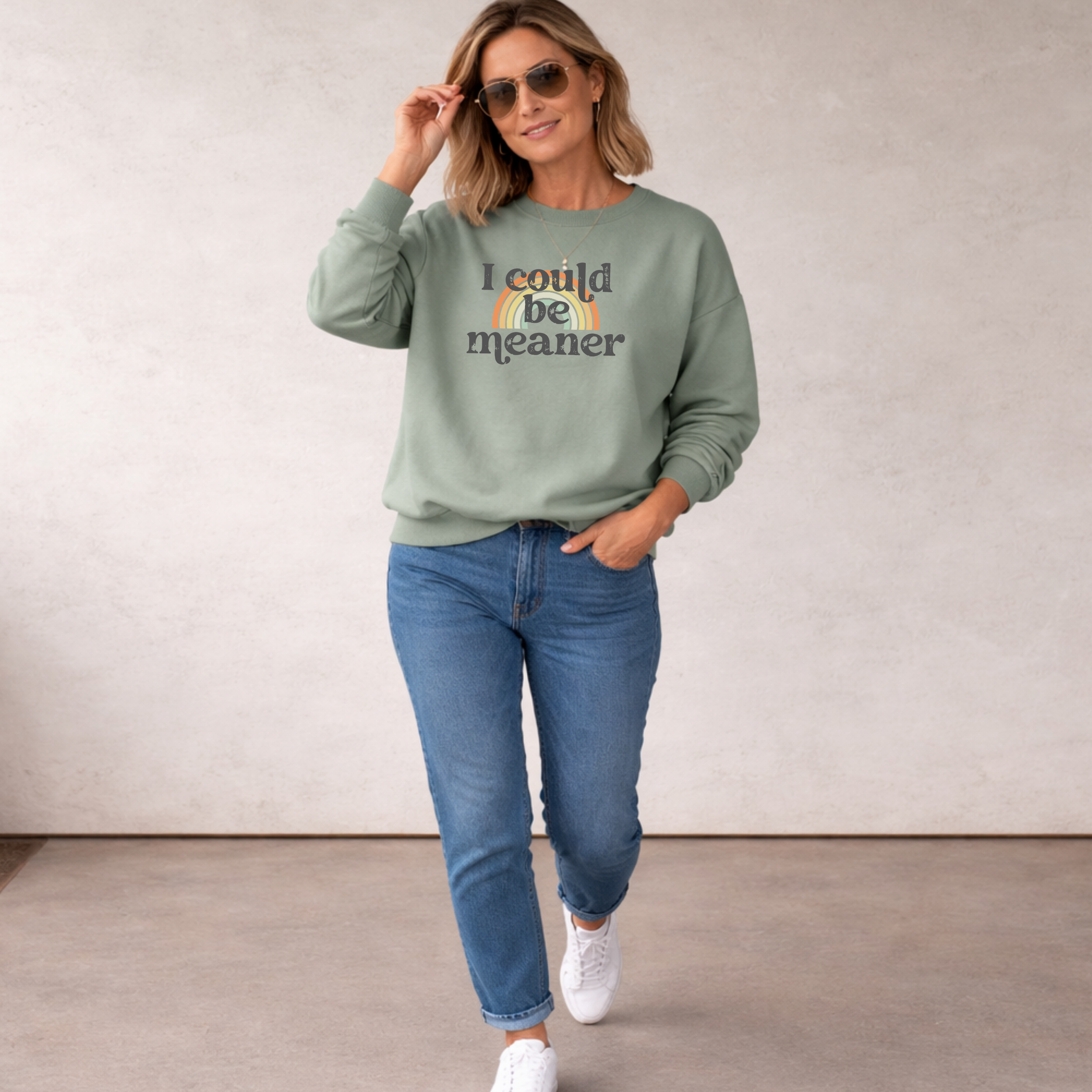 Woman wearing a green sweatshirt with a rainbow with the saying 'I could be meaner' design with jeans and sneakers.
