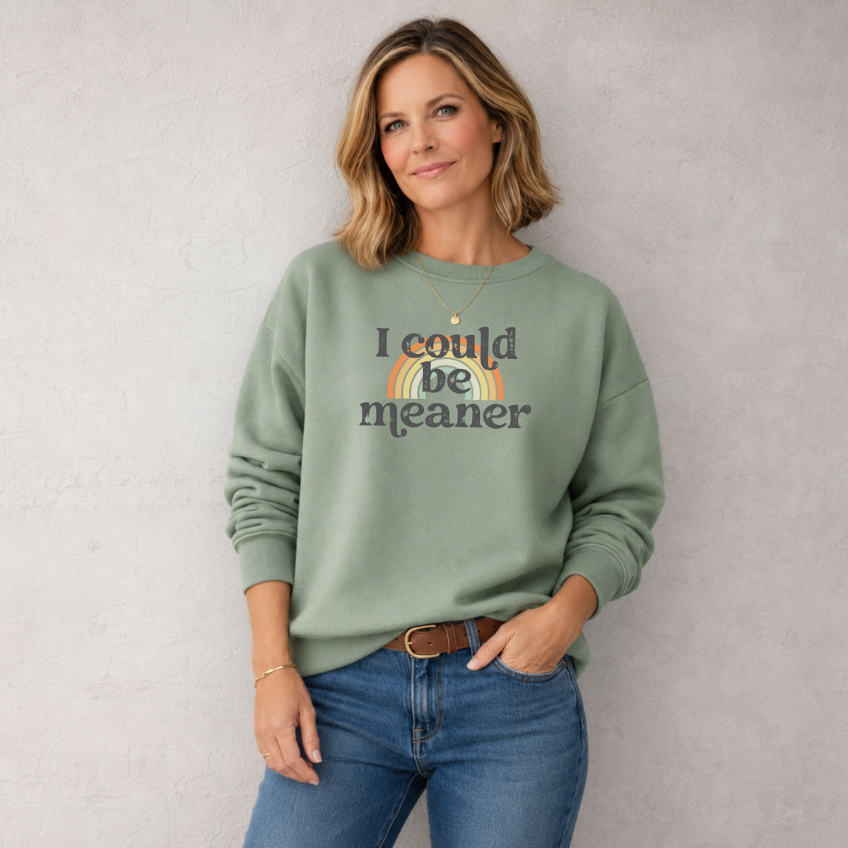Woman wearing a green sweatshirt with 'i could be meaner' text over a rainbow and blue jeans against a plain background