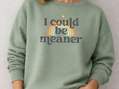 Woman wearing a green sweatshirt with 'i could be meaner' text over a rainbow and blue jeans against a plain background