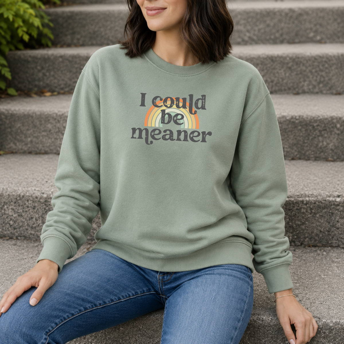 Woman wearing a green sweatshirt with 'i could be meaner' text over a rainbow and blue jeans against a plain background