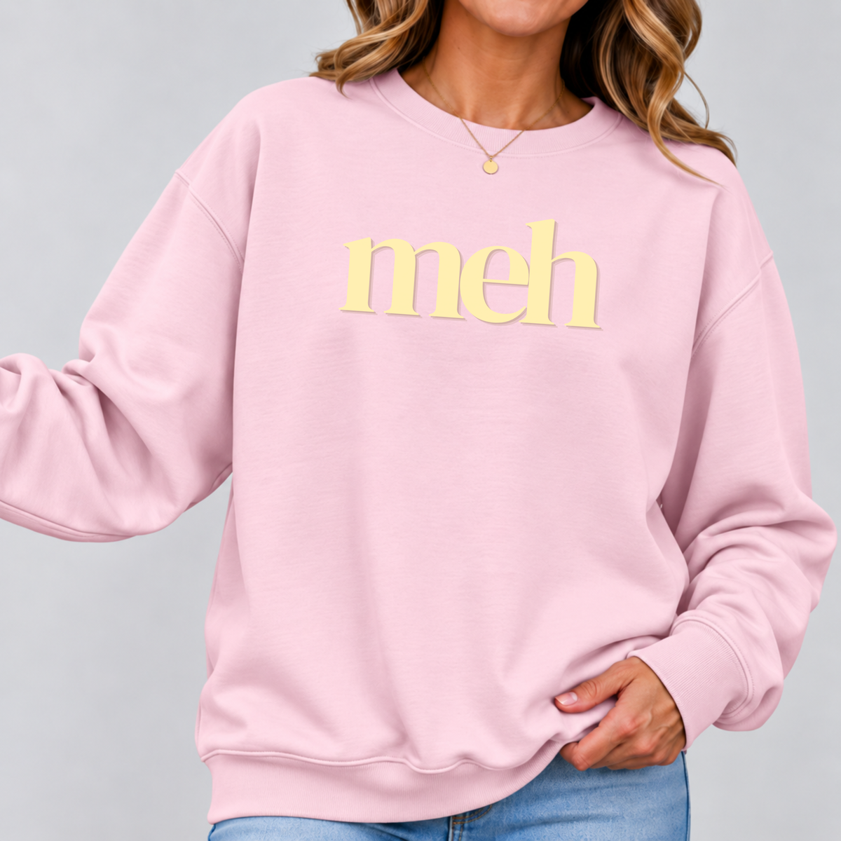 A woman wearing a light pink crewneck sweatshirt with the word 'meh' printed in yellow across the chest, paired with blue jeans against a solid gray background.