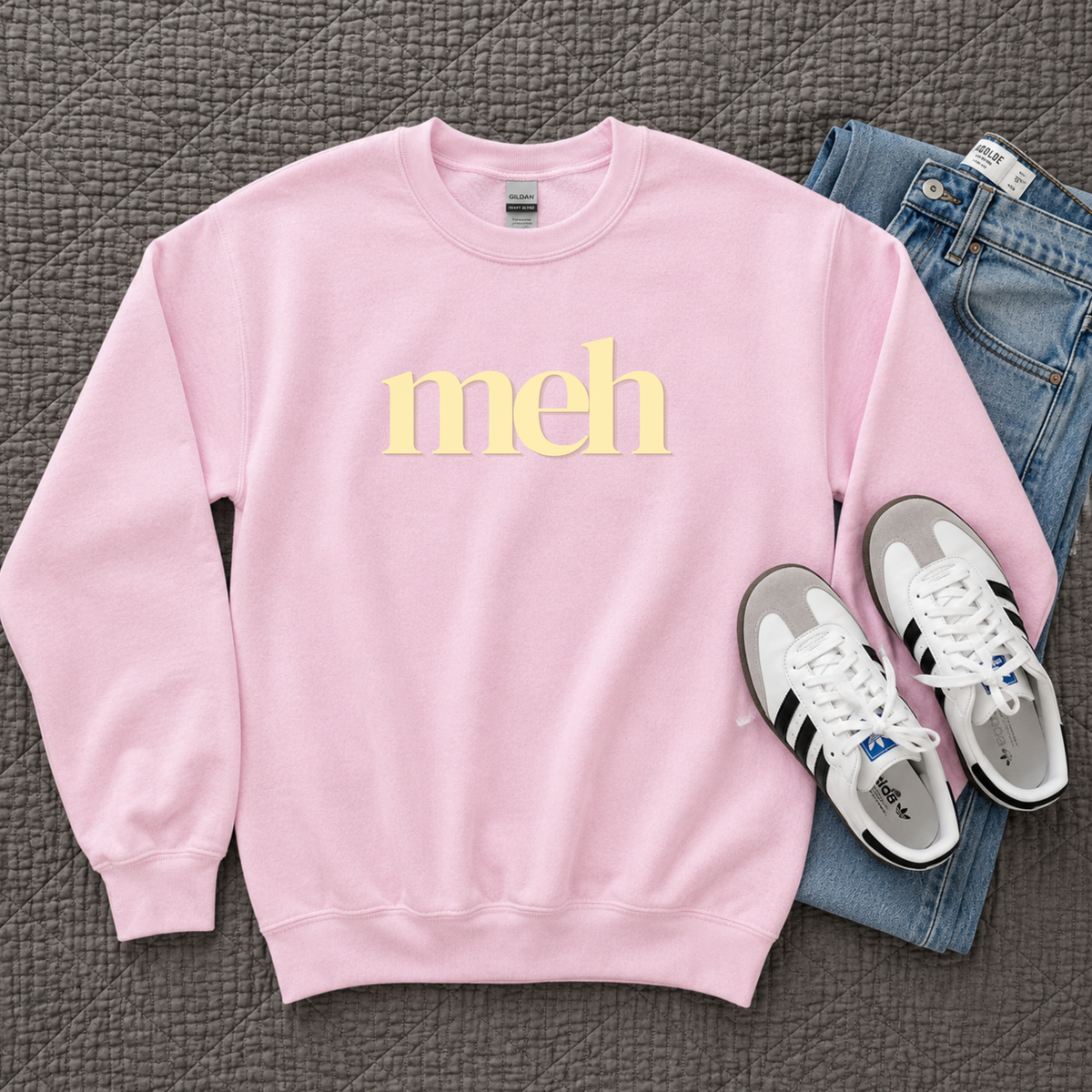 A light pink crewneck sweatshirt with the word 'meh' printed in pale yellow across the chest, styled in a flat lay with blue jeans and white sneakers on a textured gray blanket.