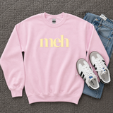 A light pink crewneck sweatshirt with the word 'meh' printed in pale yellow across the chest, styled in a flat lay with blue jeans and white sneakers on a textured gray blanket.