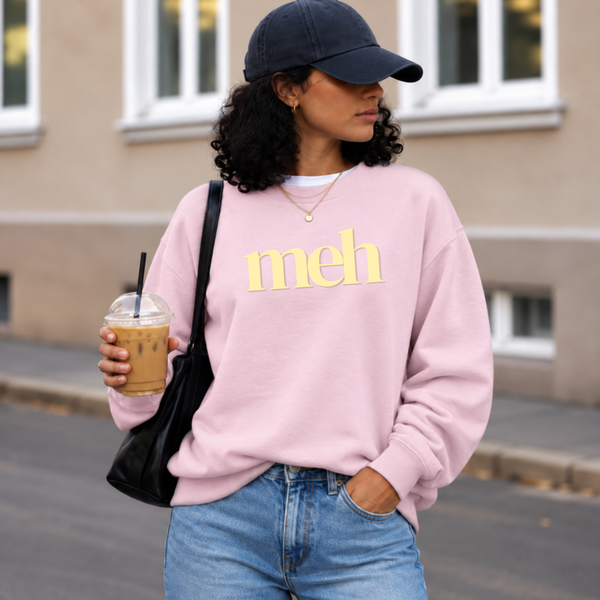 A woman wearing a light pink crewneck sweatshirt with the word 'meh' printed in yellow across the chest, paired with blue jeans, a black baseball cap, and a black shoulder bag while holding an iced coffee.