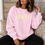 A woman wearing a light pink crewneck sweatshirt with the word 'meh' printed in yellow across the chest, sitting on a patterned couch against a rustic wood-paneled wall.