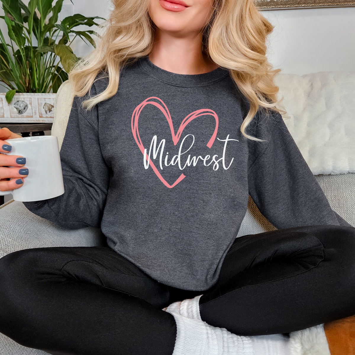 Person wearing a gray sweatshirt with 'Midwest' and a heart design, holding a mug.