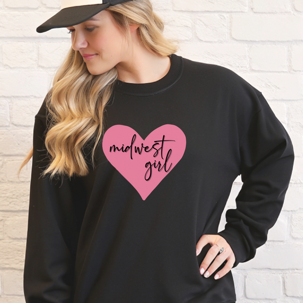 Woman wearing a black sweatshirt with a pink heart and 'midwest girl' text against a white brick wall.
