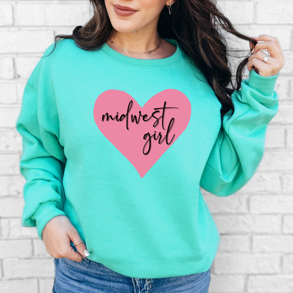 Person wearing a mint green sweatshirt with a pink heart and 'midwest girl' text against a white brick wall.