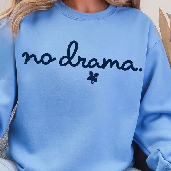 A person wearing a light blue crewneck sweatshirt with the text 'no drama.' in dark navy script and a small dark navy flower graphic underneath.