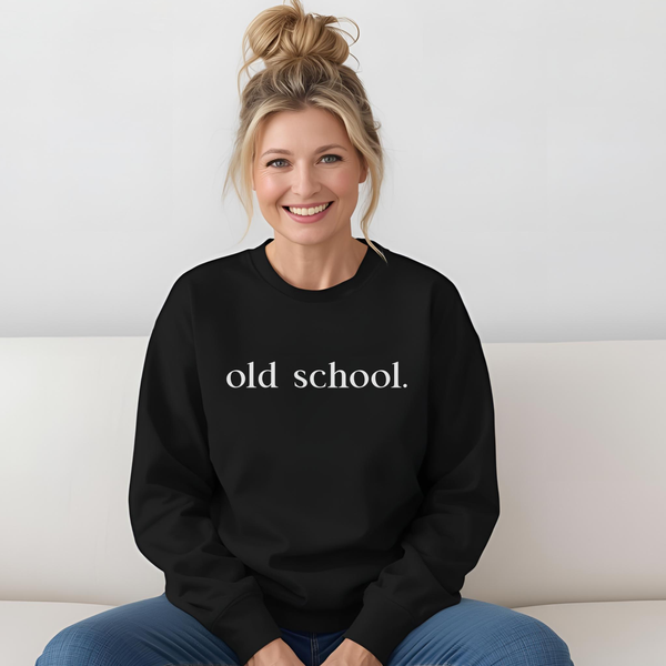 A 45-year old Woman wearing a black sweatshirt with 'old school.' white text, sitting on a white couch.