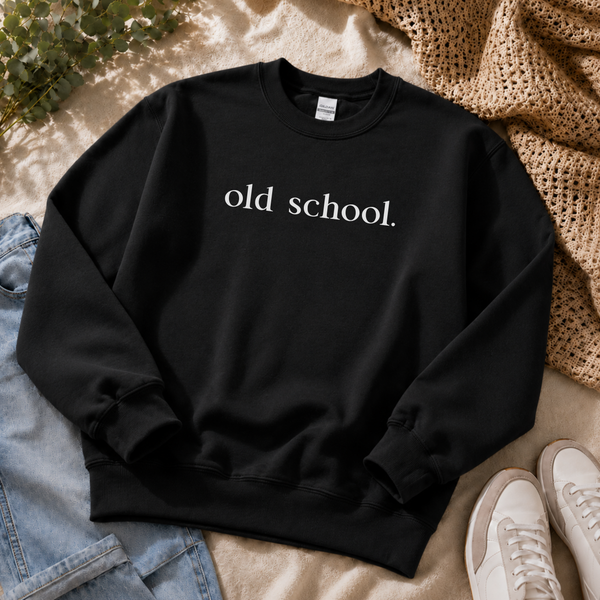 A black crewneck sweatshirt with the phrase 'old school.' printed in white serif text across the chest, styled in a flat lay with light blue jeans, white sneakers, a beige knit blanket, and green leaves on a beige fabric background.