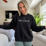 A woman with blonde hair wearing a black crewneck sweatshirt with 'old school.' printed in white text across the chest, standing in a bedroom setting.
