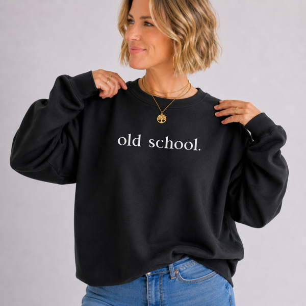 A woman wearing a black crewneck sweatshirt with the words 'old school.' printed in white across the chest, paired with blue jeans, against a light gray studio background.