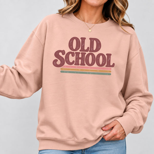 A woman wearing a dusty pink crewneck sweatshirt with a retro-style 'OLD SCHOOL' graphic in maroon and horizontal pastel stripes, paired with blue jeans against a light gray studio background.