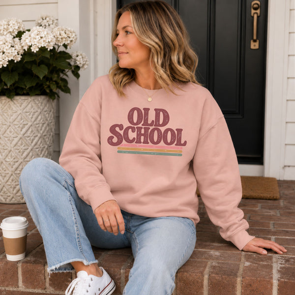 A woman sits on brick steps in front of a black front door, wearing a dusty pink sweatshirt with 'OLD SCHOOL' printed in maroon and a thin multicolor stripe graphic. She is wearing light blue jeans and white sneakers, with a coffee cup beside her.