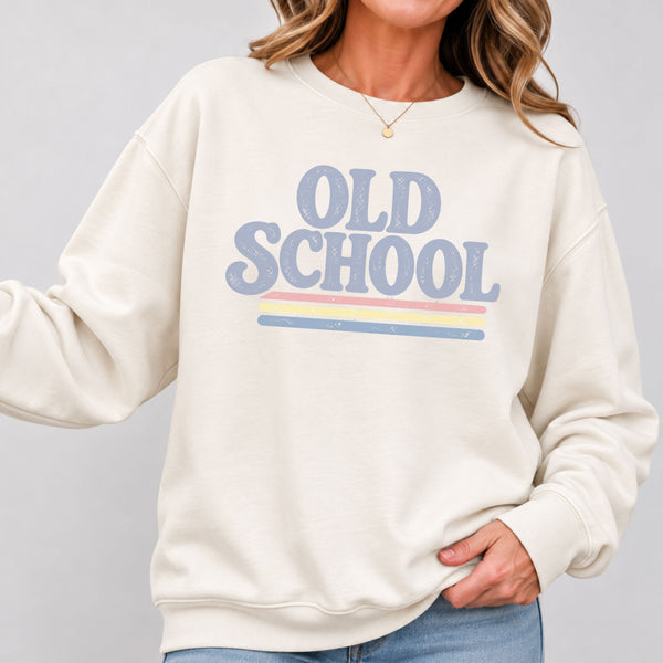 A woman wearing a cream-colored crewneck sweatshirt with a retro-style graphic that reads 'OLD SCHOOL' in dusty blue letters, accented by thin pastel stripes in pink, yellow, and blue. The photo is cropped from the chin down, showing the model wearing blue jeans against a neutral gray background.