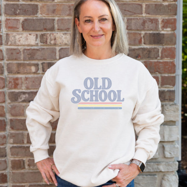 A woman with blonde hair stands in front of a brick wall wearing a cream-colored sweatshirt with a retro graphic that reads 'OLD SCHOOL' in muted blue-gray letters with pink and yellow stripes underneath.