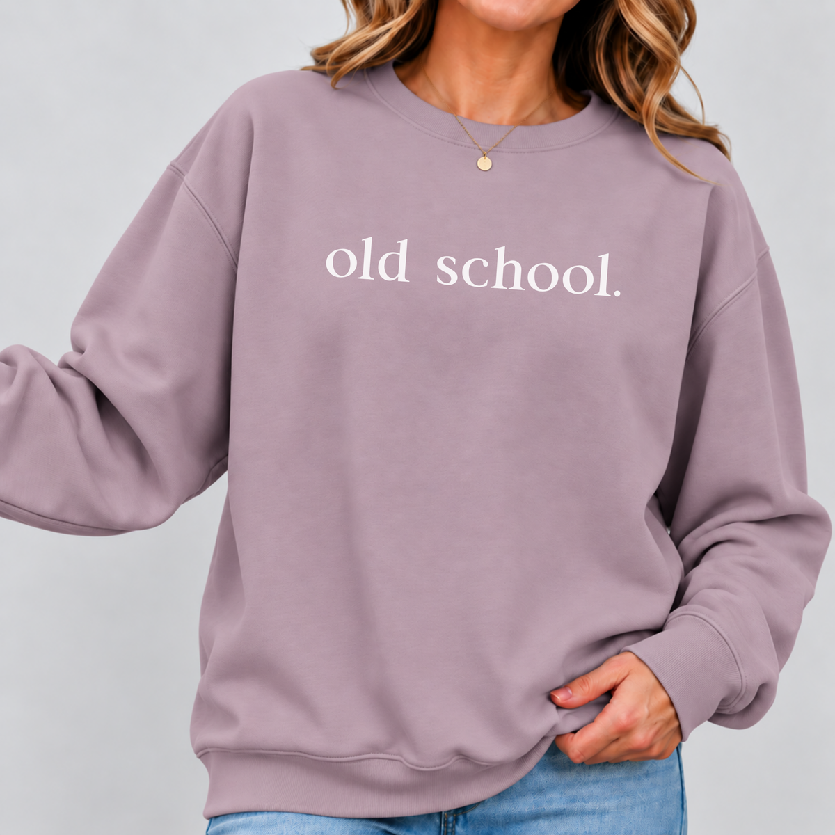A woman wearing a dusty mauve crewneck sweatshirt with the words 'old school.' printed in white across the chest, paired with blue jeans against a light gray background.