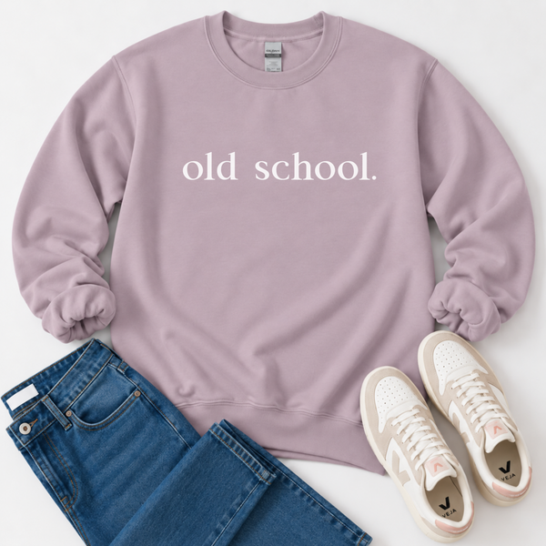 A flat-lay outfit shot featuring a dusty purple sweatshirt with the words 'old school.' printed in white across the chest, styled with blue jeans and cream sneakers on a white background.