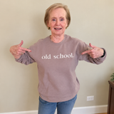 An older woman wearing a taupe sweatshirt with 'old school.' printed in white across the chest, pointing at the text with both hands.