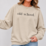 A woman wearing a beige crewneck sweatshirt with 'old school.' printed in black serif text across the chest, paired with blue jeans against a light gray studio background.