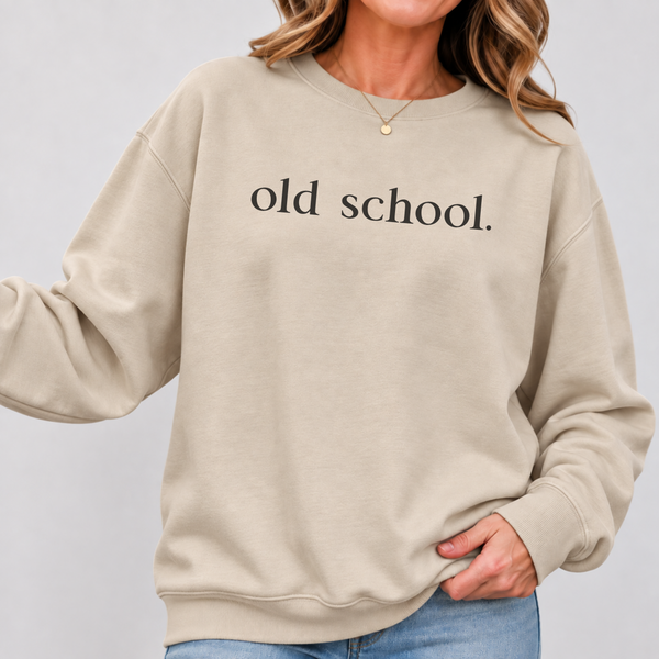 A woman wearing a beige crewneck sweatshirt with 'old school.' printed in black serif text across the chest, paired with blue jeans against a light gray studio background.